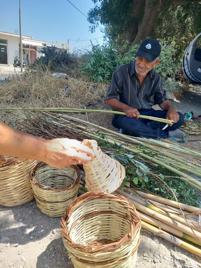 An elderly man wearing a black baseball cap and a striped shirt sits on the ground in Platani, Greece, surrounded by bundles of natural materials. He is engaged in the craft of basket weaving, with a half-finished basket in front of him and a collection of reeds and branches at his side. His hands are positioned as if he is working on the basket, demonstrating a skilled and practiced motion. The environment is outdoors, with dry vegetation, trees, and a building in the background, suggesting a warm, possibly rural or semi-rural setting. The scene captures a moment of traditional craftsmanship amidst a serene, sunlit atmosphere.