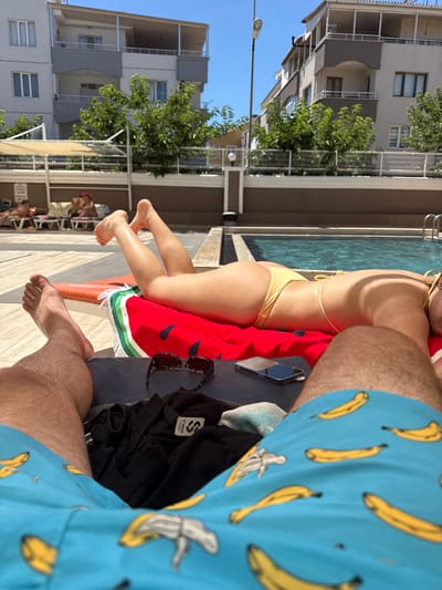 The image captures a sun-drenched scene at a poolside resort in Ayvalik, Turkey. In the foreground, the legs of two individuals are visible, suggesting they are relaxing on lounge chairs. One person's legs are in focus, wearing bright blue swim trunks adorned with a banana print, while the other person's legs are also visible, suggesting they are lying next to each other.

Behind them, a woman in a yellow bikini is sunbathing on a red towel with a watermelon pattern. She is lying on her stomach, her legs propped up. A smartphone and a pair of sunglasses are placed on a dark surface near her.

The background reveals the turquoise water of a swimming pool, with a ladder visible on the right side, indicating the pool's edge. Beyond the pool, a fence separates the pool area from a brown-toned wall, which then leads to a multi-story building with balconies and windows. Several trees with green foliage are situated between the pool area and the building. In the distance, other lounge chairs with people relaxing can be seen under a shaded canopy, and another building is visible, suggesting a larger resort complex. The sky is clear and blue, indicating fair weather.