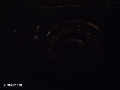 The image shows a dark, close-up view of a decorative metallic object. The object appears to be circular with a Greek key or meander pattern embossed around its circumference. The pattern is highlighted with a reddish-orange glow, suggesting it might be illuminated or made of a material that reflects light in that way. The rest of the object and its surroundings are extremely dark, making it difficult to discern further details about its form or context. The text "HONOR 400" is visible in white at the bottom left corner of the image, likely indicating the device used to capture the photograph. There are no people, actions, or interactions visible in the image. The location in the city of Kumkurgan, Uzbekistan, is not discernible from the visual information provided.