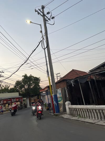 The image depicts a street scene in Karangtanjung, Indonesia, likely during the late afternoon or early evening, as indicated by the warm, soft light. The sky is a pale gradient of blue and orange, suggesting sunset or sunrise. The perspective is angled, with a prominent utility pole and a tangle of electrical wires dominating the left side of the frame. A street lamp on the pole is illuminated, casting a bright glow.
On the right side of the image, a street runs alongside buildings. The architecture appears to be a mix of traditional and modern, with a tiled roof on one structure and a more contemporary facade on another. There are several motorbikes on the road, with some people riding them, indicating a busy street. One red motorbike in the foreground is clearly visible. The overall atmosphere is that of an active urban environment transitioning into dusk.
Several signs with text are partially visible. One sign, partially obscured by a tree, appears to be colorful and possibly advertising. Another sign, more prominent on the lower right, has bold text that reads "SLIC 1B" and some smaller text that includes prices like "12,000". The presence of these signs, combined with the motorbikes and the general street activity, points to a commercial area. The weather appears calm, with no visible rain or strong winds. The image captures a slice of everyday life in this Indonesian locale.