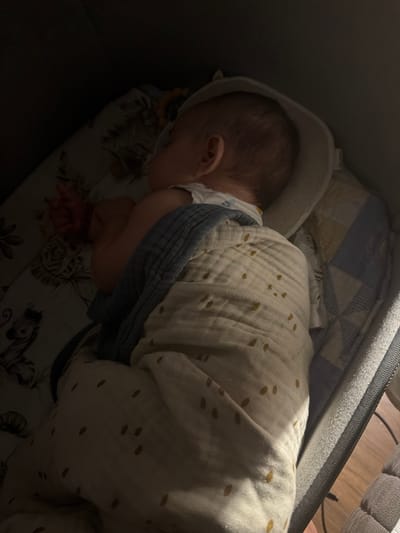 A newborn infant is sleeping in a bassinet in Pontonx-sur-l'Adour, France. The infant is swaddled in a light-colored muslin blanket with scattered mustard-yellow dots and is wearing a white onesie. A blue-grey textured blanket is partially visible under the muslin blanket. The bassinet has a padded rim and patterned sheets featuring animals and floral motifs on the left side, and a quilted pattern of blue and yellow squares on the right. The overall lighting is dim, with a light source illuminating the infant from the right side, casting shadows. The scene appears to be indoors, likely in a bedroom.