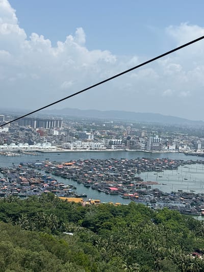 The image captures a panoramic view of Nanwan Village, China, a coastal settlement characterized by its dense network of floating structures and boats. The foreground features a lush, green hillside, presumably part of the natural landscape surrounding the village. Beyond the hillside, the village itself unfolds, with a vast expanse of the sea occupied by numerous floating houses and fish farms. These structures are densely packed, forming a vibrant, almost chaotic, mosaic of rooftops and fishing nets. Many of the floating homes appear to be constructed from wood and corrugated metal, with some painted in various colors, including red and orange. A multitude of boats, ranging from small fishing vessels to larger cargo ships, are docked or navigating the waterways between the floating structures.

In the mid-ground, a more developed part of the village is visible, featuring modern white buildings with clean architectural lines, possibly representing commercial or residential areas. A prominent green-domed structure, likely a religious building or landmark, stands out amidst the urban landscape. Further in the background, a city skyline emerges, with tall, multi-story buildings indicating a more urbanized section of the region. The distant horizon is marked by rolling hills or mountains, partially obscured by atmospheric haze. The sky above is a pale blue, dotted with fluffy white clouds, suggesting a clear, sunny day. A thick black cable, likely from a cable car or power line, diagonally traverses the upper portion of the image, intruding slightly into the otherwise natural and man-made landscape. No individuals are clearly discernible in the image. The scene depicts a bustling waterfront community, highlighting the intersection of traditional fishing practices and modern development.