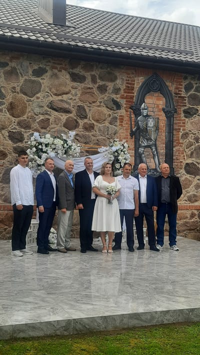 A wedding party of nine people stands on a tiered, marble-like platform in front of a stone and brick wall with a mural of a knight. The individuals are positioned from left to right: a young man in a white button-down shirt and black pants; a man in a dark blue suit; an older man in a gray suit and blue shirt; a man in a black suit; the bride in a white dress with puffy sleeves and holding a small bouquet of white flowers; a man in a white button-down shirt and dark pants; a man in a dark blue suit and dark pants; and a man in a black jacket and dark jeans. The bride is standing in the center of the group. The backdrop features floral arrangements of white roses and greenery. The mural of the knight is framed by an arched gothic structure. The environment appears to be outdoors, with a glimpse of green grass in the foreground. The location is identified as Жодзішкаўскі сельскі Савет, Belarus.