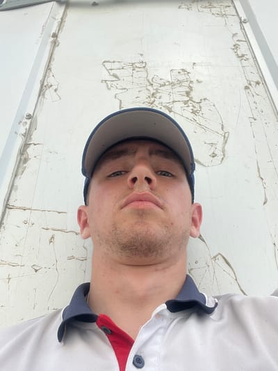 A low-angle, close-up selfie of a young man wearing a blue and gray baseball cap and a white polo shirt with navy and red trim. He is looking directly at the camera with a neutral expression. The background is a rough, white textured surface with some dirt or debris. The image appears to be taken outdoors in a somewhat industrial or utilitarian setting. No specific identifiable landmarks for Аснежыцкі сельскі Савет are visible.