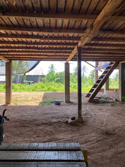The image depicts the interior of an unfinished wooden structure in Buketdalam, Indonesia. The foreground features a rough-hewn wooden bench, with a dark, possibly metal object to its left. The floor of the structure is composed of packed dirt. Concrete pillars support the wooden ceiling and upper structure. To the right, a wooden staircase leads upwards. The background reveals lush green vegetation, including banana trees and palm trees, indicating a tropical environment. Beyond the vegetation, there are glimpses of houses with corrugated metal roofs. The overall impression is of a work in progress in a rural or semi-rural setting.