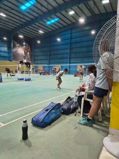 The image depicts an indoor badminton court in Phagwara, India. The court is illuminated by overhead lights, and the walls are lined with blue corrugated metal. A large banner featuring a badminton player is displayed on the back wall. Several individuals are present on and around the court. In the foreground, two large blue badminton bags and a water bottle are placed on the floor. To the right, a large industrial fan partially obscures the view of a woman sitting on a white chair, wearing a floral print top and dark shorts. Another person, wearing a light blue shirt and dark shorts, is also seated nearby, with their legs crossed. On the court, several badminton players are engaged in matches, including a player in a black t-shirt and shorts on the left, and a player in a grey t-shirt and shorts in the center. An umpire is seated on a raised chair, observing the game. Additional spectators are visible in the background. The court surface is a green synthetic material with white boundary lines.