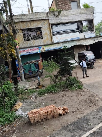 The image depicts a street scene in Benapole, Bangladesh, on an overcast day. In the foreground, a pile of bricks sits on muddy ground. A narrow, overgrown ditch runs alongside the road. A standing water pipe with a spigot is visible. Further back, a three-wheeled auto-rickshaw is parked on the pavement. A man wearing a light blue shirt and gray pants stands beside it, looking to the right, with a black backpack on his left shoulder. Behind the auto-rickshaw, a building with multiple stories and shops on the ground floor is visible. Signs in Bengali are displayed on the building's facade. Trees and foliage surround the area, with some growing on the building's roof. The overall environment appears somewhat disheveled and in need of maintenance.