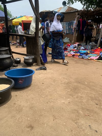The image depicts a bustling outdoor market scene in Nigeria, likely during the daytime with the sun casting shadows. In the foreground, a woman wearing a white hijab and a blue patterned dress stands in a dirt-floored market. She is looking towards the viewer and holding a small red object, possibly fruit, in her hand. Beside her, several large metal cooking pots and a large blue plastic basin are visible, suggesting food preparation or sale.
In the background, a stall is laden with a colorful assortment of clothing, with items neatly folded or hung. More clothing is visible in piles to the left of the stall, creating a vibrant and busy atmosphere. Several other people are present in the market, including individuals appearing to be vendors or shoppers interacting with the goods. One person in the background is wearing a white shirt and dark trousers, seemingly engaged in an activity related to the stall. A yellow awning with "MAGGI" branding is prominently displayed on one of the stalls, indicating a food product brand. The overall scene conveys a sense of lively commerce and daily life in a Nigerian market.