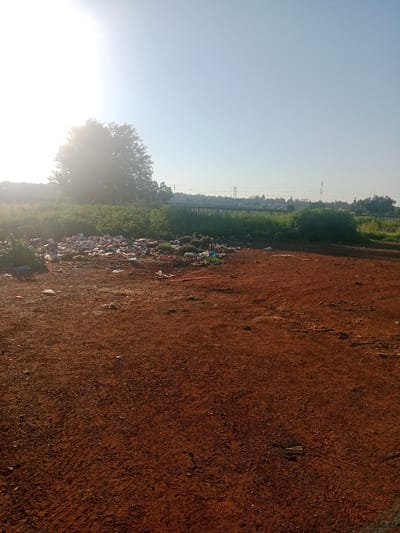 An outdoor scene in Soweto, South Africa, depicts an unpaved, reddish-brown dirt field in the foreground, showing visible tire tracks and scattered small debris. In the midground, to the left, a substantial accumulation of refuse, consisting of various plastic items and discarded materials, is present. Behind this waste, a strip of green vegetation, including bushes and tall grass, runs horizontally across the frame. Further back, a dark fence or barrier separates this area from distant structures, likely residential, and more green foliage. The background features a clear, light blue sky. A bright, intense sun in the upper left quadrant causes significant glare, partially obscuring a large, leafy tree. In the far distance, multiple electrical transmission towers are visible against the horizon. No individuals or active subjects are present.
