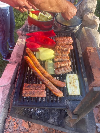 In Javorje, Montenegro, an outdoor barbecue is underway on a grill. Two people are visible; one person's hands are in the frame, holding a fork over a pot of red and yellow bell peppers. Another person's arms and torso are also partially visible. On the grill, several sausages and pieces of meat are cooking, along with slices of zucchini and bell peppers. The grill is made of black metal and rests on a structure of rough stones and bricks. The surrounding area is covered in dry grass and scattered red brick pieces. In the background, a metal pot with a glass lid and more stone formations are present.