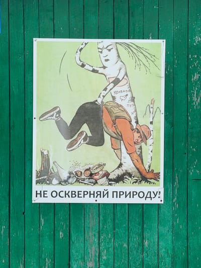 A poster depicts a person in mid-air, leaping away from a collection of trash that is forming a monstrous shape. The person, wearing a hat, orange shirt, dark pants, and sneakers, has a backpack and is reaching out with their right hand. The trash pile includes cans, bottles, and a gas mask, suggesting pollution. The background is a light green, representing nature, with a stylized tree. Text in Cyrillic runs vertically along the right side of the poster. The poster is attached to a weathered, green wooden fence. The scene is likely intended to convey a message about environmental protection, specifically urging people not to pollute nature. The person's expression and pose suggest alarm or disgust at the pollution.