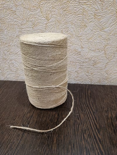A spool of light brown jute twine sits on a dark brown wooden surface. The twine is wound tightly around the spool, creating a cylindrical shape. A loose end of the twine extends from the spool and lies on the wooden surface, trailing to the left. The background is a textured, pale yellow wall with a raised, organic pattern, possibly wallpaper. The lighting appears to be even, suggesting an indoor setting, and there is no indication of the time of day or weather conditions. No people are visible, and there is no text in the image. The scene is simple and focuses on the object and its immediate surroundings, with a shallow depth of field that blurs the background slightly. The overall impression is one of a utilitarian object placed in a domestic or workshop setting.