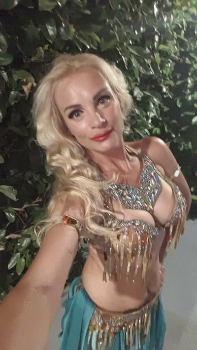 A medium shot captures a blonde woman in a bejeweled, revealing belly dancing costume, with her arm outstretched towards the camera, creating a selfie-like pose. She is positioned in front of a dense, dark green foliage wall, likely outdoors at night, as indicated by the lighting. The woman has bright red lipstick and her eyes are looking directly into the camera. The costume is ornate, featuring numerous rhinestones and dangling gold-colored elements. The lower half of the image shows a teal-colored skirt adorned with a matching embellished belt. The overall impression is one of a performance or celebratory setting. No specific location in Efeler, Turkey, can be identified from the visual information alone.