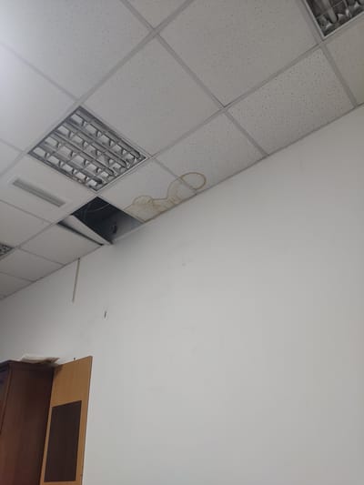 The image shows an interior view of a room with a dropped ceiling. A fluorescent light fixture is visible in the upper left quadrant, and a section of the ceiling has been removed, exposing wires and the space above. There is a water stain on the ceiling tiles to the right of the removed section. The walls are painted white, and part of a wooden cabinet and door are visible in the lower left corner. The scene appears to be within a building in Egizbulak, Uzbekistan.