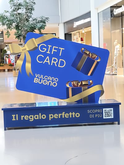 The image shows a large blue "GIFT CARD" display with a golden bow and gift box illustrations. Below the gift card, a blue stand with yellow text reads "Il regalo perfetto" (The perfect gift) and "SCOPRI DI PIÙ" (Discover more) alongside a QR code. The display is situated in the common area of a shopping mall, likely in the city of Nola, Italy, given the text "VULCANO BUONO" prominently displayed on the gift card, which is the name of a well-known shopping mall in Nola. The background shows shops and artificial trees typical of an indoor shopping environment. No people are visible in the immediate foreground or around the display.