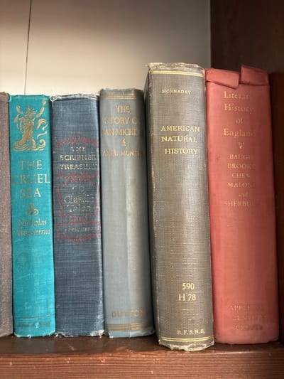 The image displays a forensic close-up of seven hardbound books arranged on a dark brown wooden shelf. The books exhibit varying states of wear, indicating age. No individuals are present, and no actions or interactions are depicted.

From left to right, the visible book spines are:
1.  A partially visible, plain light grey or brown book.
2.  A teal-colored book titled "THE CRUEL SEA" by "Nicholas Monsarrat," featuring golden embossed designs, including a mythical figure at the top and a Celtic knot near the bottom. The publisher "Alfred A. Knopf" is visible at the base.
3.  A dark blue-grey book titled "THE SCRIBNER TREASURY OF CLASSIC TALES," with embossed red-gold text and noticeable fraying along its top and bottom edges. "SCRIBNER" is printed at the base.
4.  A light blue-grey book titled "THE STORY OF SAN MICHELE" by "AXEL MUNTHE," with "DUTTON" at the base.
5.  A grey-tan book titled "AMERICAN NATURAL HISTORY" by "HORNADAY," showing the call number "590 H 78" and "R.F.S.N.S." at the base. The top and bottom edges of its spine are frayed.
6.  A reddish-orange book titled "Literary History of England," listing "BAUGH BROOKE CHEW MALONE and SHERBURN" as authors, with "APPLETON CENTURY CROFTS" at the base. The top of this spine is significantly torn.
7.  A partially visible dark brown book on the far right.

The books rest on a wooden shelf, which shows minor imperfections. Above the books, a plain, light-colored surface, likely a wall or the back of the bookshelf, is visible. The specific location within Monterey, United States, cannot be determined from the visual information provided in the image.