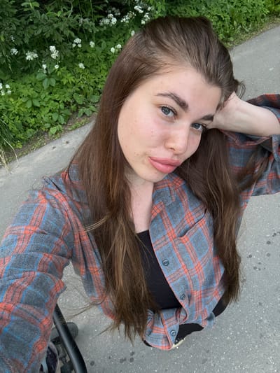 A young woman with long, brown hair is captured in a selfie taken outdoors. She is wearing a grey, red, and blue plaid shirt over a black top. Her left arm is resting on her head. The background consists of lush green foliage with small white flowers, and a grey paved surface, likely a sidewalk or path. The image is focused on the woman's face and upper body. The location is specified as Shevlyagino, Russia.