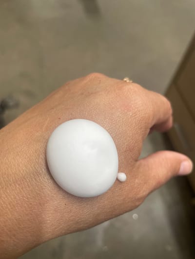 A close-up, overhead shot shows a person's left hand with a large dollop of white cream on the back of it. A tiny bead of the same cream is visible to the right of the larger dollop. The hand is resting on a textured, neutral-colored surface. A hint of a ring is visible on one of the fingers, as is a portion of a cardboard box in the background. The image was taken in the city of Tervolovo, Russia.