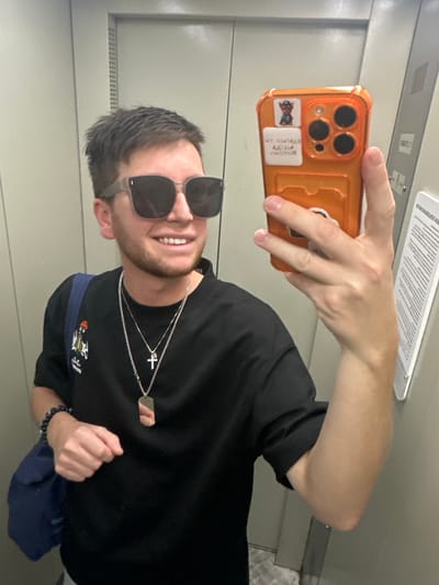 In an indoor setting, a young man is taking a selfie. He is wearing a black t-shirt, dark sunglasses, and multiple necklaces, one of which features a cross pendant and a dog tag. He also has a beaded bracelet on his right wrist. His phone, which has an orange case with three cameras, is held in his right hand. On the phone case, there are two small stickers. The background appears to be the interior of an elevator, with grey metallic walls and a document or sign attached to the ceiling. The lighting suggests it is daytime. The man is smiling, indicating a positive emotion. The location is identified as Tokaryovo, Russia.