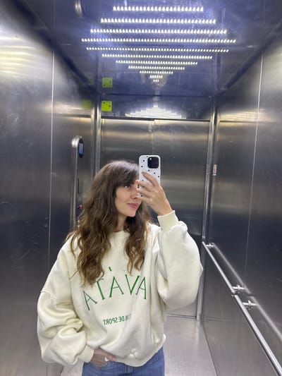 A young woman with long, wavy brown hair is taking a selfie in an elevator. She is wearing a cream-colored sweatshirt with green lettering that says "ATAVA" and "CLUB DE SPORT" in reverse. Her right hand is holding her phone, which has a white case with black polka dots, and her left hand is resting on her hip. The elevator has stainless steel walls and ceiling, with a row of bright LED lights on the ceiling. The floor appears to be metallic as well. The elevator is located in Tnuvot, Israel.