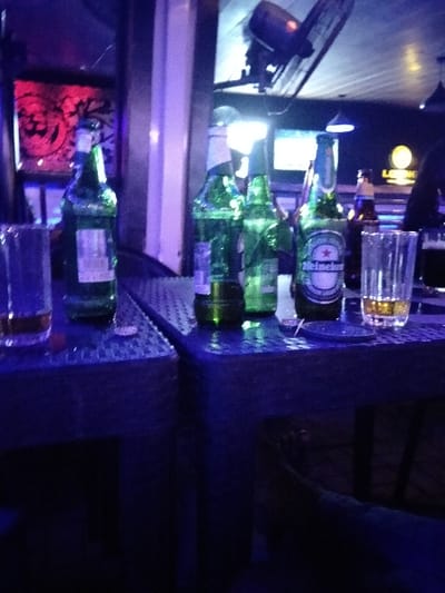Several empty and partially filled green glass beer bottles stand on two dark blue, possibly metal, tables. Next to the bottles are two glasses containing remnants of beer. The lighting is dim, with some ambient light from a bar or nightclub setting. In the background, there are additional bottles, glasses, and lighting fixtures, suggesting a bar environment. A fan or vent is mounted on the ceiling. The atmosphere is relaxed and informal.