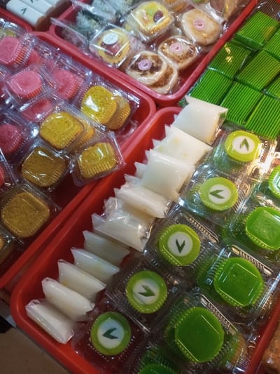 The scene displays a close-up, overhead view of various individually packaged food items, likely desserts or snacks, arranged within red plastic trays. This setting is observed in Lê Thanh Nghị Ward, Vietnam. No individuals are present in the frame.
The items are meticulously organized into distinct rows and groups by color and shape. In the upper-left, partially visible, are white cylindrical items. Adjacent to these, within a red tray, are multiple clear plastic containers each holding a square-shaped cake; some are vibrant yellow, while others are pinkish-red. Further to the upper-right, another red tray contains diverse small cakes or pastries, individually sealed in clear plastic. These include square items with visible fruit toppings and round items featuring a spiral design.
Dominating the right side of the image are several rows of green-hued food items. One section features uniform rectangular, layered green cakes, each encased in clear plastic. Below these, a row of small, opaque white items, some with a faint yellow tint, are individually sealed in clear plastic pouches. Parallel to these white pouches are rows of round, green-rimmed cakes with a white center adorned by a small green leaf-like design, each in its own clear plastic container. Finally, a section displays rows of bright, solid green, square-shaped gelatinous cakes, also individually presented in clear plastic containers. The overall impression is an organized display of confectionery or prepared foods for retail.