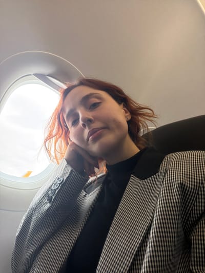 A female individual with auburn hair is seated inside an aircraft cabin, positioned in a window seat. She is depicted in a medium close-up, gazing directly towards the camera. Her right hand is positioned under her chin, with fingers curled. She wears a black turtleneck garment under a black and white checkered blazer featuring black lapels. A small silver stud earring is visible in her right ear. To her left, an aircraft window (porthole) shows an overcast sky and a blurred yellow aircraft tail fin, consistent with Pegasus Airlines. The white overhead panel and cabin ceiling are visible above her. The location is an aircraft in flight or on the ground at or near Sabiha Gökçen International Airport (SAW), which is situated in the vicinity of Tuzla, Istanbul, Turkey.