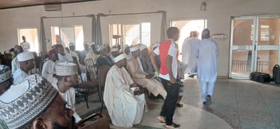The image depicts an indoor gathering of men, likely at a community event or a religious service, in Sabon Gari North, Nigeria. The setting appears to be a hall with a light-colored floor and walls. Natural light streams in from windows and doorways, suggesting it is daytime.

The subjects are predominantly men, many of whom are wearing traditional attire, including white robes and caps of various styles, common in many Muslim communities. Some individuals are seated, while others are standing or in the process of moving. One person, in the foreground, is seen from behind wearing a red and white t-shirt and black track pants, suggesting a more casual presence or perhaps staff.

In the background, there are doorways leading to other areas, and large glass doors that appear to lead outside. The overall atmosphere seems orderly and communal. The presence of chairs arranged in rows indicates a planned assembly. Two dark bags, resembling duffel bags or briefcases, are visible near the glass doors, possibly belonging to attendees.

The image does not contain any visible text. The lighting and attire suggest a warm, possibly humid day, typical of Nigeria. The gathering appears to be a moment of shared experience, with people engaged in conversation or listening, indicative of a planned event or meeting.