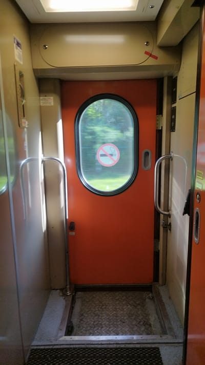 The image shows the interior of a train car, likely a vestibule area between cars. A bright orange door with an oval window dominates the center of the frame. A "no smoking" symbol is visible inside the window. To the left of the orange door, there is a gray metallic wall with a vertical handle. A silver grab bar extends from this wall. To the right of the orange door, another door, partially open and appearing to be a lighter color, reveals a metallic handle. The floor is a combination of diamond-plate metal and a dark mat. The lighting suggests an overcast day outside, casting soft light into the train car. There are no people visible in the image. The environment appears utilitarian and functional, typical of public transportation.
