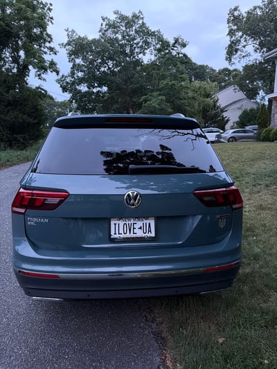The image shows the rear view of a teal Volkswagen Tiguan SEL parked on an asphalt driveway. The car has a New York license plate that reads "I LOVE UA". A Ukrainian trident emblem is visible on the right side of the rear bumper. The car is parked in front of a grassy yard with trees and a house in the background. The sky is overcast. The location is identified as Flanders, United States of America.