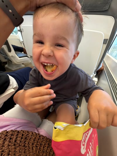 A young child, with fair skin and light brown hair, is being held up in what appears to be a train carriage. The child is smiling broadly, with their mouth open and a piece of snack food inside. They are wearing a dark grey t-shirt and shorts. Their hands are holding a bag of chips, and one hand is reaching into the bag. The child's head is being gently held by an adult's hand, wearing a dark watch on their wrist. In the background, there are white train seats and a window. The scene does not provide any specific visual cues to indicate the location is the city of Tarragona, Spain.