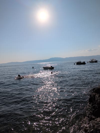 This is a sunny, outdoor, eye-level shot of the Adriatic Sea near Kraljevica, Croatia. The sun is bright and high in the sky, casting a glare on the water. Several small boats are anchored offshore. In the foreground, a person is swimming with an inflatable tube. The water is dark blue and calm, with sunlight glinting off its surface. Rocky shoreline is visible on the right side of the frame. In the background, a range of mountains is visible on the horizon. The sky is clear and blue.
