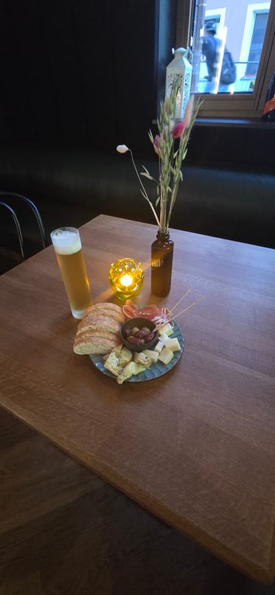 This image captures a cozy scene on a wooden table, likely in an indoor setting with low lighting, suggesting evening or a dimly lit room. The primary focus is a charcuterie board, artfully arranged with cubes of cheese, cured meat, olives, artichoke hearts, and slices of bread. Beside the board sits a tall glass of beer, its foam head creating a frothy contrast with the golden liquid. A small, amber-colored glass bottle holds a bouquet of dried flowers and grasses, adding a rustic touch to the display. A decorative, yellow glass candle holder glows warmly, casting a soft light on its surroundings and contributing to the intimate atmosphere. In the background, a white lantern with intricate cutouts is visible on a ledge, and beyond that, a window reveals a glimpse of the outside, though details are blurred. The overall impression is one of a relaxed gathering or a quiet moment of indulgence, hinting at a comfortable ambiance. The setting appears to be a restaurant or a home in Bregenz, Austria, given the location context. There is no visible text in the image.