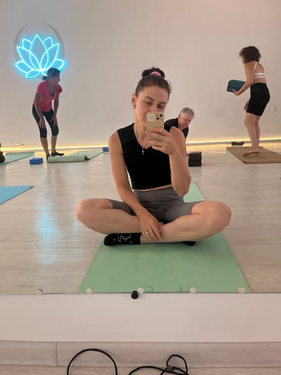 A woman in a black crop top and grey shorts is sitting in a cross-legged pose on a green yoga mat. She is holding a smartphone in her right hand, taking a selfie. She is wearing socks with a white design. Her left hand rests on her knee. In the background, another woman in a white sports bra and black shorts is holding two green yoga blocks. To the left of that woman, another person is bent over. Further to the left, a woman in a pink t-shirt and black capri pants is also bent over. A neon lotus flower light is visible on the wall behind them. The floor is made of light-colored wood. The scene appears to be a yoga studio. The image does not provide any information about the location being in Aksu, Kazakhstan.
