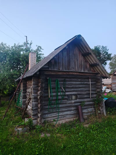 The image depicts a small, rustic wooden log cabin situated in a grassy yard. The cabin has a steeply pitched roof covered with corrugated material. A brick chimney extends from the roof on the left side. Several green garden hoses are coiled and hung on the side of the cabin, along with some rope. A metal pipe stands vertically near the right corner of the cabin, and a small window with a patterned glass is visible. The surrounding environment is overgrown with grass, with trees visible in the background to the left and right. There are some miscellaneous items stacked to the right of the cabin, including what appear to be wooden planks and some fabric. The overall scene suggests a rural setting.