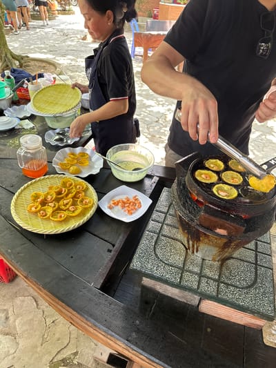 The image captures a street food scene in Tân Thạch, Vietnam, showcasing the preparation and serving of a local delicacy. A man is seen using tongs to remove small, round, golden-yellow pancakes with a shrimp topping from a specialized stove with multiple concave molds. The stove, heavily used and blackened from cooking, sits atop a gray tiled surface on a dark wooden table.
Next to the stove, a woman is focused on preparing the finished dishes. Several plates and a woven tray hold the cooked pancakes, while condiments and dipping sauces are neatly arranged. The scene suggests an outdoor setting with a casual ambiance, likely a roadside eatery popular among locals and tourists. A few blurry figures can be seen in the background, potentially other customers or passersby. The overall composition highlights the culinary practice and informal food culture typical of the region.