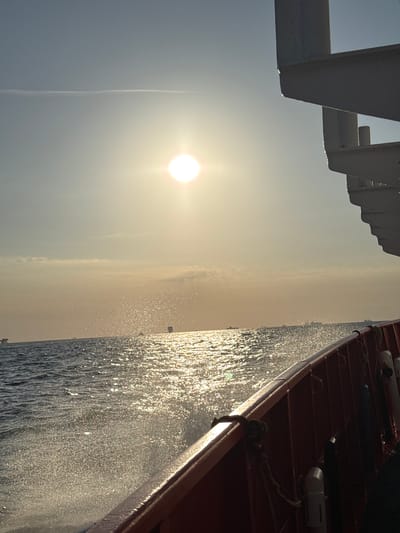 The image captures a sun-drenched scene at sea, likely near Marina di Ravenna, Italy, as suggested by the context. The bright sun, positioned high in the sky, casts a brilliant glare on the choppy water. The water itself is alive with spray and foam, indicating movement, possibly from the vessel on which the photograph is taken. In the distance, several ships and possibly oil rigs are visible on the horizon, suggesting a busy maritime area. The foreground is dominated by the side of a red vessel, with visible railings and ropes, implying the perspective is from a moving boat. The sky is a pale blue, with a faint cloud streak and the sun's intense light creating a hazy effect. The overall impression is one of a bright, active day at sea.