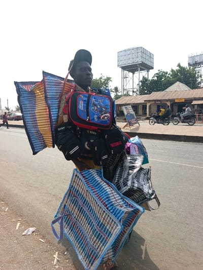 This image depicts a street vendor in Nassarawan Eggon, Nigeria, heavily laden with a variety of goods for sale under a bright, clear sky.
The central subject is a young man, wearing a dark baseball cap, looking intently towards his right. He is almost completely obscured by the sheer volume of items he is carrying. On his chest, he displays a vibrant blue and red child's lunchbox or small backpack featuring Captain America and the text "Captain America" and "Avengers." Attached around his mid-section are several black pouches or small bags. Visible text on these includes "Balenciaga," "OFF White" (likely brand replicas), and partially "STORY TO." Large, colorful woven bags, patterned in blue, white, red, and black stripes and checks, are strapped to his back and hang down to his sides and front, creating a striking silhouette. A small red tag with an "RB" logo in a circle can be seen on one of the lower blue and white bags.
The vendor is standing on the edge of a paved road, which shows some scattered debris. In the background to the right, a row of modest, single-story buildings with corrugated iron roofs suggests shops or local businesses. Two motorcycles, each with a rider, are visible on the road in front of these establishments. A political billboard displaying a man's face and some text stands near the buildings. Further back, green trees and two prominent, elevated metal water towers with square reservoirs stand against the bright sky. On the far left, a person walks in the distance, and a car is parked. The scene captures a typical bustling street environment, highlighting the daily life and commerce of the area.