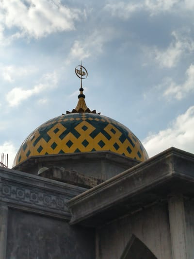 The image shows the dome of a mosque located in Blangbitra, Indonesia. The dome is yellow with a geometric pattern of green diamonds. At the top of the dome is a golden spire topped with the Arabic inscription of Allah. The dome is situated atop a gray building structure. The sky in the background is light blue with scattered white clouds.