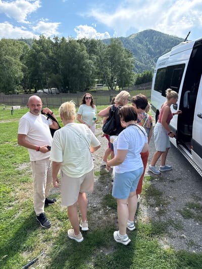 A group of seven people, four women and three men, are gathered near the open door of a white minibus on a sunny day. The minibus appears to be parked on a gravel surface with green grass and some trees in the background. In the far background, a mountain range covered with trees is visible under a blue sky with white clouds.

The group is casually dressed, suggesting a tour or excursion. One man on the left, wearing a white t-shirt and beige pants, is holding a black bag strap in his hand. He has a serious expression on his face. To his right, a woman with blonde hair, wearing a light-colored t-shirt and beige shorts, is facing away from the camera. Another woman, in a light green t-shirt and pink shorts, stands behind her, holding a water bottle.

Further to the right, a woman with dark hair, wearing a black top and a patterned shirt, is partially visible. Next to her, a woman in a white t-shirt and light blue shorts is also facing away. In the foreground, a woman in a pink tank top and a light blue denim skirt is reaching into the open door of the minibus, holding a dark object in her hand. Another woman, in a red tank top and white shorts, stands by the door.

The environment suggests a rural or semi-rural setting, possibly a stop on a tour route in Kupchegen, Russia. The presence of the minibus and the gathering of people indicates a planned activity.