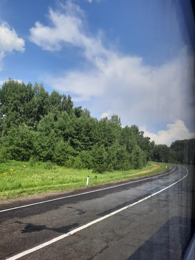 The image depicts a curved asphalt road with white lane markings. The road appears wet, with visible reflections of the sky and surrounding environment. On the left side of the road, there is a grassy embankment dotted with small white flowers and some yellow wildflowers. Beyond the embankment, a dense line of green trees forms a backdrop. The sky above is blue with scattered white cumulus clouds, and a darker, more diffused cloud formation is visible in the distance on the right. The scene is captured from a moving vehicle, as indicated by the slight motion blur and the reflection of what appears to be a window frame on the far right of the image. There are no people or animals visible in the frame. This scene is typical of a rural or semi-rural roadside environment, possibly on the outskirts of the city of Krapivinskiy, Russia, during daylight hours.