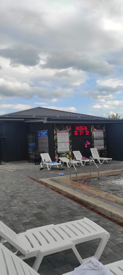 The image depicts an outdoor recreational area, possibly a spa or water park, in Tulskiy, Russia. Several white plastic lounge chairs are arranged on a paved patio surrounding a body of water, which appears to be a pool or hot tub with a grated edge. The water is not clearly visible, but its texture suggests movement or bubbling.
A dark building with a sloped roof forms the backdrop. Mounted on the building's exterior wall is a digital display showing red LED numbers: "30 006.26" and "36 37 32". Above this display are informational posters with floral decorations. A blue sign with an icon of a horseshoe and text in Cyrillic is also visible on the building.
The weather appears to be partly cloudy, with large, soft white clouds dominating the sky. Sunlight filters through the clouds, suggesting it is daytime, possibly late morning or afternoon. The overall atmosphere seems relaxed and inviting, with the lounge chairs indicating a place for patrons to rest and enjoy the facilities. There are no people actively using the water or chairs in the frame, but there are a few towels and a pair of blue flip-flops visible on the chairs and ground, implying recent use.