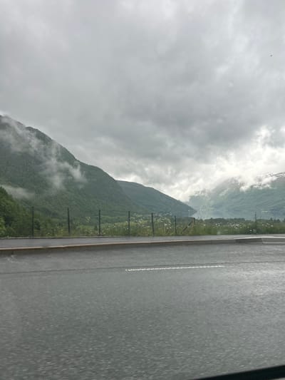 The image depicts a scenic view of a misty mountain landscape, likely taken from the side of a road in Røldal, Norway. The primary subjects are the lush green mountains, partially obscured by swirling clouds and mist. A paved road runs along the right side of the frame, marked with a white line. Along the edge of the road, a wire fence with vertical posts is visible, separating the road from the mountainside. The mountains are densely covered with trees, and patches of white, possibly snow or rock, can be seen higher up on the slopes. The sky is overcast with heavy, gray clouds, suggesting a cloudy or rainy day. The overall mood is serene and perhaps a bit dramatic due to the mist and the imposing mountains. There is no visible text in the image. The lighting suggests it is daytime, though the overcast conditions create a subdued atmosphere. The scene evokes a sense of natural beauty and the ruggedness of the Norwegian terrain.