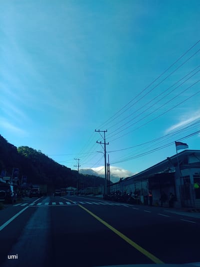 The image depicts a road in the city of Dolat Rayat, Indonesia, with a clear blue sky overhead. A paved road runs through the center of the frame, with a yellow line dividing the lanes. On the left side of the road, a steep, tree-covered hill rises, partially obscured by shadows. On the right side, a building with a weathered facade is visible, and an Indonesian flag is flown from a pole on its roof. Several utility poles with power lines run parallel to the road. In front of the building, a row of parked motorcycles and a few people are present. A crosswalk is marked on the road, and several vehicles, including cars and trucks, are either parked or driving on the road. The lighting suggests it is daytime, possibly early morning or late afternoon, given the long shadows. The word "umi" is visible in the lower left corner.