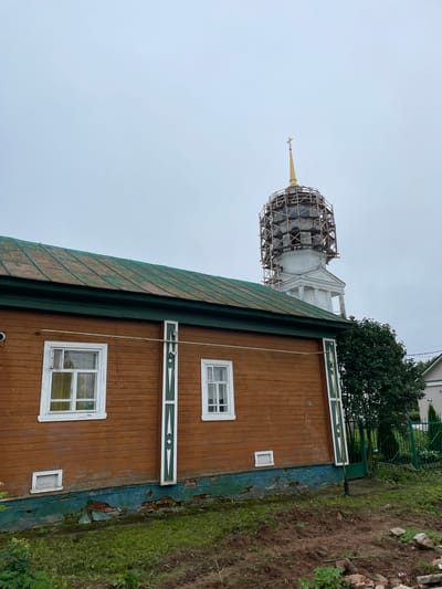 In the city of Kstynino, Russia, a wooden house with a weathered green roof stands in the foreground. The house is painted brown and features white-trimmed windows. The foundation of the house is visible, showing exposed brickwork and peeling green paint. To the right of the house, a tall white structure, possibly a church bell tower, is partially obscured by scaffolding. A golden cross tops the tower. A mature tree with dark green foliage grows next to the tower, and a green metal fence encloses a garden area with a white house in the background. The sky is overcast, casting a muted light over the scene. The ground in front of the house is uneven, with patches of grass and disturbed soil, suggesting recent construction or landscaping work.