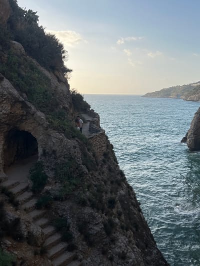 This image depicts a picturesque coastal scene with a rocky cliff face on the left and the sea on the right. A stone pathway winds along the cliff, leading to an arched opening that appears to be a small cave or tunnel. Steps carved into the rock descend from the pathway towards the cave entrance. A couple, a man and a woman, stand on the pathway, appearing to be looking towards the sea. The sea is a deep blue-green, with gentle waves. In the distance, a headland covered in vegetation juts out into the water. The sky above is a pale blue with a few wispy clouds, and the sun appears to be setting or rising, casting a soft, warm light. The location is identified as being in Trappeto, Italy, suggesting it might be part of a coastal path or a scenic viewpoint in that area.