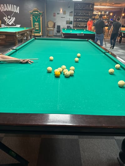 The image depicts a billiards game in progress in a bar or game room. A pool table dominates the scene, covered in green felt and scattered with multiple white balls and one yellow ball. A person's hand rests on the table next to a cue stick, suggesting an imminent shot.

In the background, other people are present, moving around in the bar area. A well-stocked bar is visible, featuring shelves filled with various bottles. The branding "Piramida" is displayed on a wall near another pool table.

There is no specific evidence to confirm the location as San Vicente Del Raspeig/Sant Vicent Del Raspeig, Spain, and therefore the location cannot be verified with certainty.