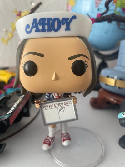 The image shows a close-up of a Funko Pop figurine of Robin Buckley from Stranger Things, wearing a white sailor hat with "AHOY" written in blue. She is holding a whiteboard with "YOU RULE" and "YOU SUCK" written on it, with tally marks under "YOU SUCK." The figurine is standing on a clear plastic base. In the background, out of focus, are various objects including a globe, what appears to be another toy, and part of a racing car model. The overall setting is likely an indoor display or shelf, given the arrangement of objects and the lighting. The image does not provide any information about the city of Brovary, Ukraine, or its specific location within it.
