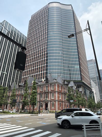 The image depicts a street scene in Chiyoda, Japan, featuring a prominent modern skyscraper with a curved facade and a reddish-brown grid pattern, alongside a more classical brick building with ornate architectural details and steeply pitched roofs. In the foreground, a white SUV is stopped at a crosswalk. Traffic lights are visible, with one red light illuminated. A black van is also present on the road. A street lamp with a speaker attached hangs over the intersection. The sky is overcast. Trees line the sidewalks of the brick building.