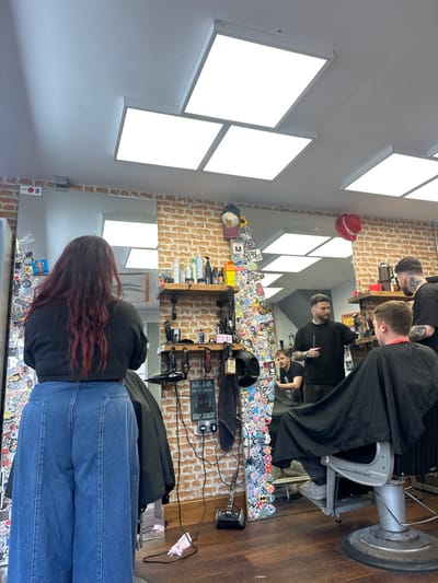 The image depicts the interior of a barbershop, identified as being located in Salisbury, United Kingdom.
The scene features a well-lit space with a white ceiling containing multiple large, rectangular LED panel lights. The walls are partially covered with exposed brickwork, above which are smoother, lighter-colored sections. A significant portion of one wall and a large mirror frame are extensively decorated with numerous colorful stickers. The floor is dark wood.
In the foreground, a person with long, dark, wavy hair with red undertones is seen from behind, wearing a black long-sleeved top and wide-leg blue denim jeans. They are seated in a black barber chair, mostly obscured by a black cape.
Reflected in the large wall mirror to the right, a male barber with dark hair, a beard, and visible neck and arm tattoos is observed standing. He wears a black t-shirt and holds a black tool, appearing to attend to a male client seated in a barber chair. The client has short, light-colored hair and is also draped in a black cape. To the left of this primary interaction, another male individual with short light hair and a black t-shirt is partially visible in the reflection, seated or leaning. Further to the right in the reflection, another male barber or staff member is partially visible, also in a black t-shirt.
Along the brick wall, above the mirror's lower edge, are two wooden shelves displaying various hair products in bottles and jars. Below these shelves, barber tools such as clippers and a hair dryer are mounted on the wall. A black motorcycle helmet rests on a lower shelf or counter. Visible electrical cords extend from wall sockets to various appliances. On the floor in the foreground, near the center, a small black stick-style vacuum cleaner and a white medical face mask are present. A framed sign reading "hello sweet cheeks HAVE A SEAT" is affixed to the brick wall.